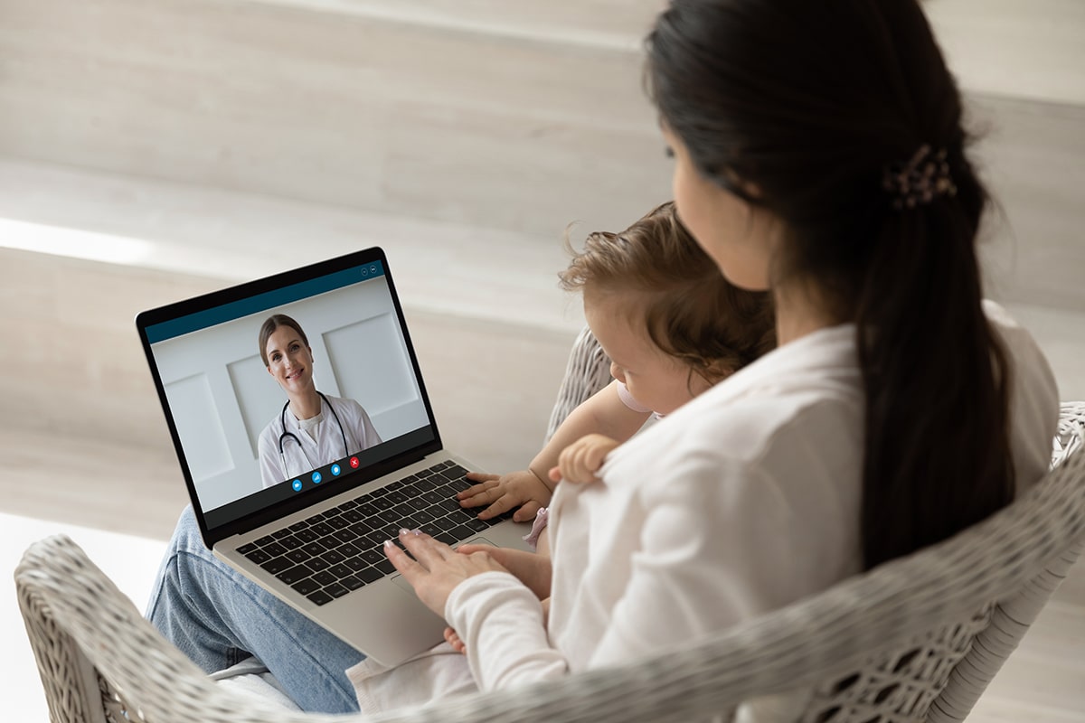 Loving mother hugging holding baby little daughter using laptop making video call to female pediatrician at home