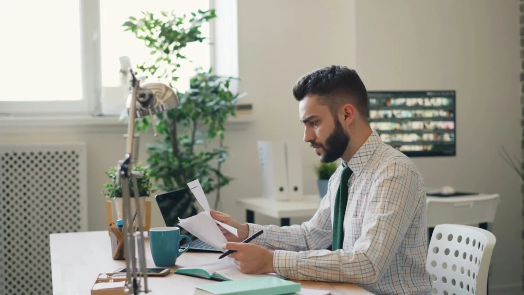 Man in a checked shirt and tie reviewing documents at a desk with a laptop, coffee cup, and plants, in a modern office setting, reflecting themes of health insurance and business planning.