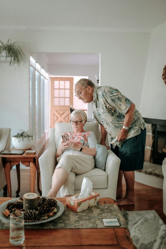 Elderly couple in a cozy living room, woman sitting on a chair using a smartphone, man leaning over to assist, emphasizing support and connection in managing health conditions.