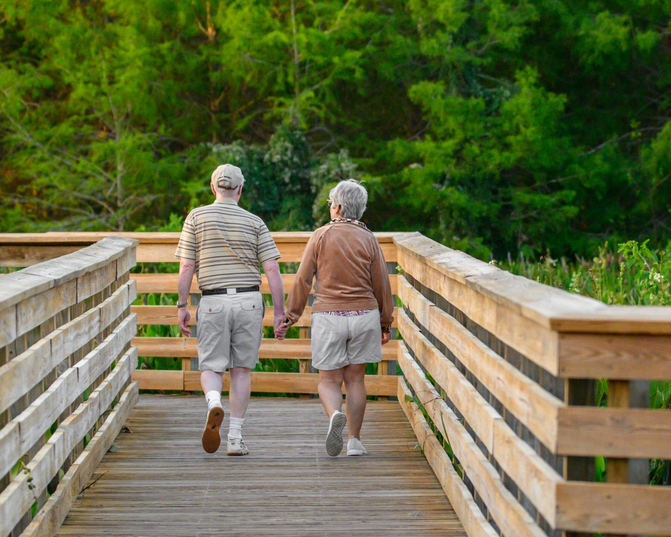Elderly couple walking hand-in-hand on a wooden boardwalk, surrounded by lush greenery, symbolizing active lifestyle and companionship in retirement.