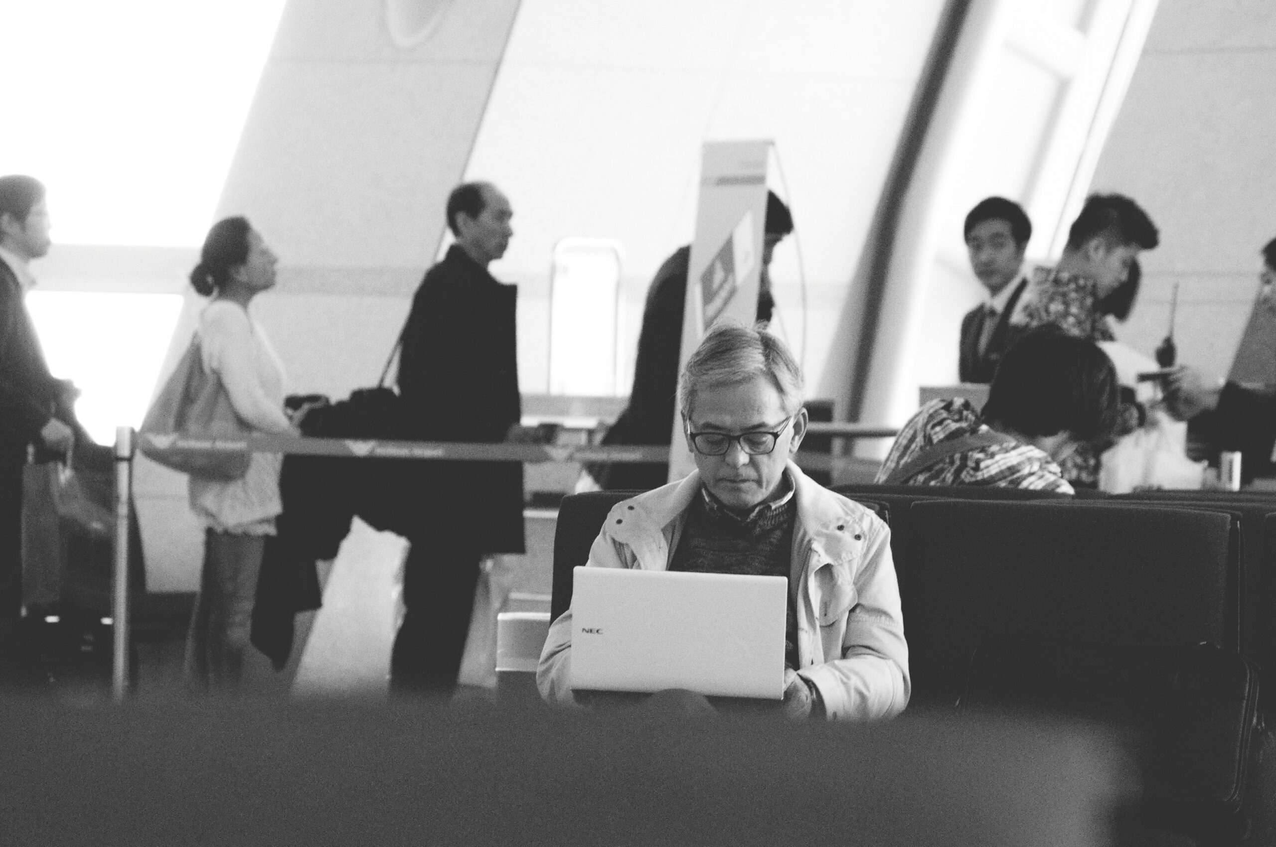 Person using a laptop in an airport lounge, surrounded by travelers, illustrating the theme of planning and decision-making for Medicare enrollment and eligibility.