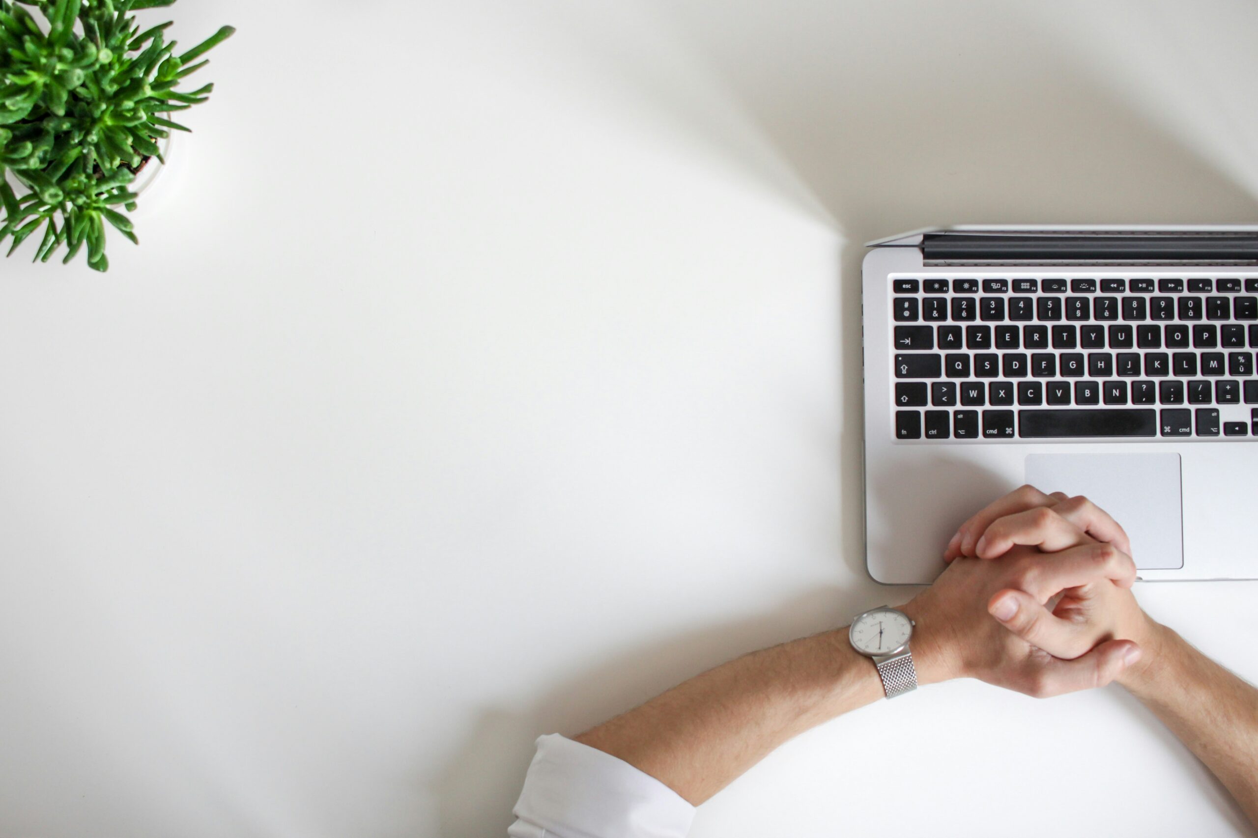 Hands clasped on a desk beside a laptop and a small potted plant, symbolizing strategic planning and decision-making related to health insurance options.