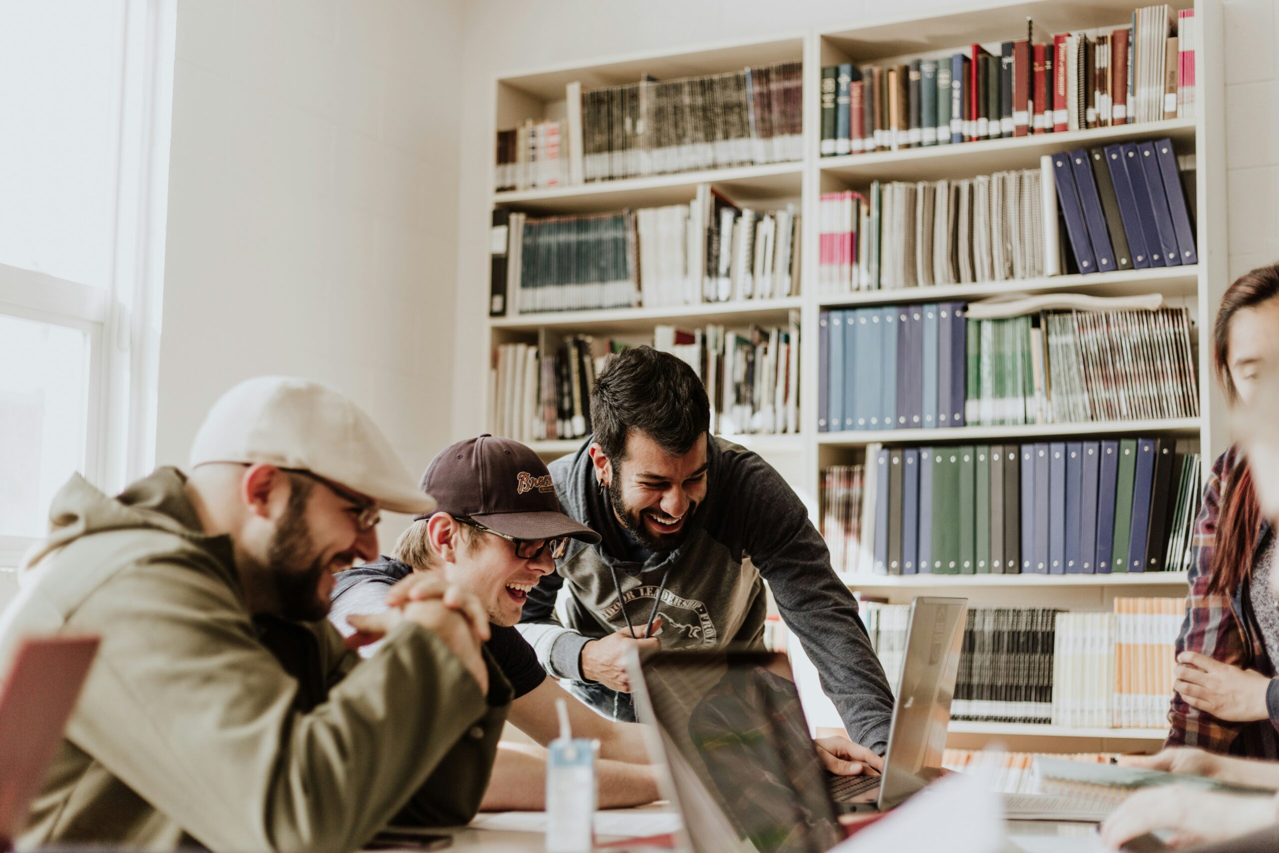Group of young professionals collaborating and laughing around a laptop in a library setting, emphasizing teamwork and engagement in strategic planning for group health insurance options.