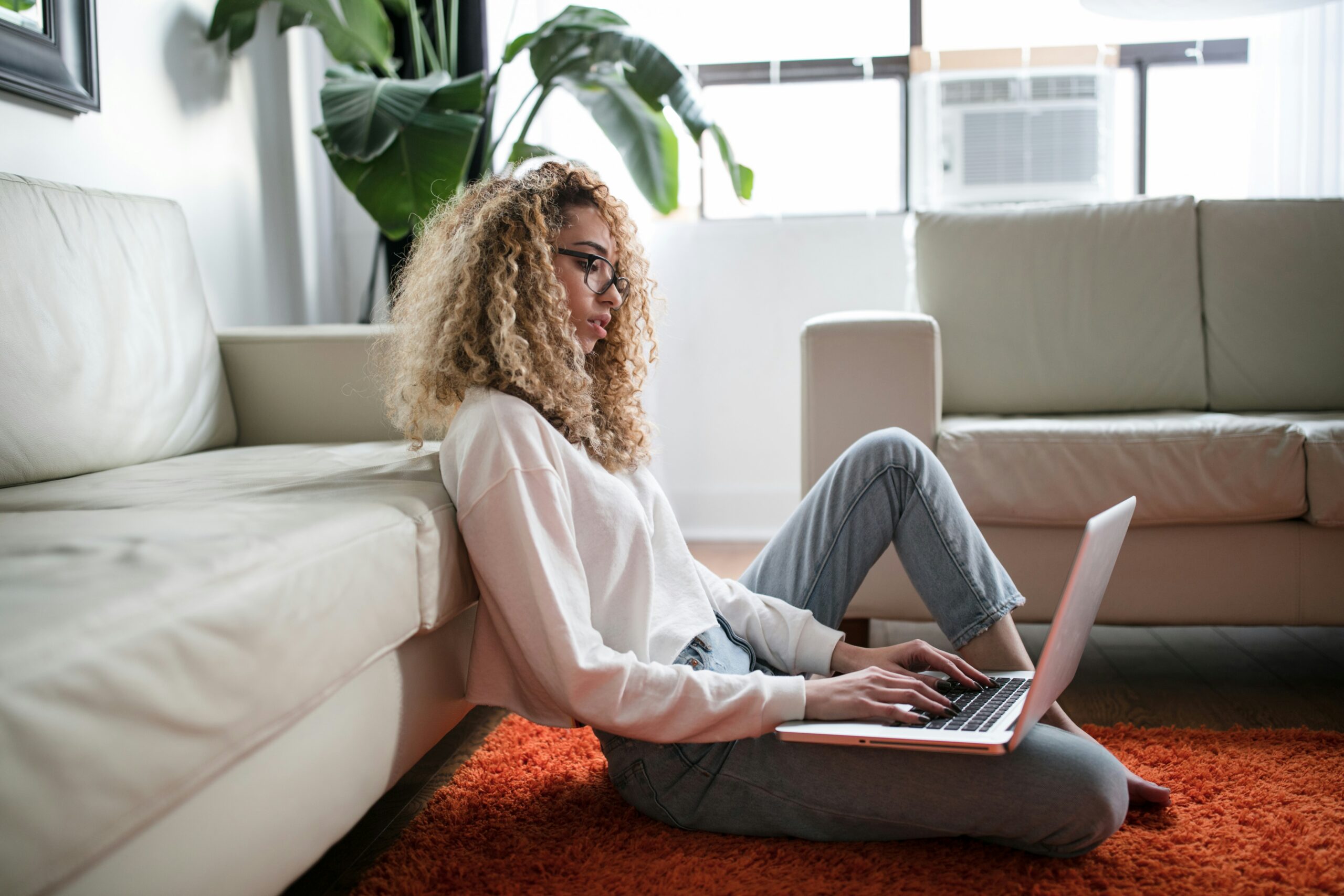 Young woman with curly hair working on a laptop while seated on an orange rug in a modern living room, emphasizing the importance of technology in managing ACA enrollment and insurance options in Florida.