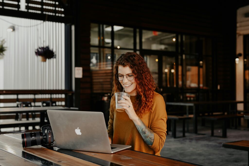 Woman with curly hair and glasses smiling while using a laptop in a casual workspace, holding a drink, suggesting a relaxed environment for freelancers and gig workers exploring health insurance options in Florida.
