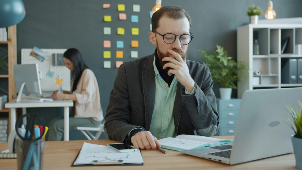 Man in a blazer and glasses contemplating at a desk with a laptop, notebook, and financial documents, while a woman works at a computer in the background, illustrating a professional environment related to health insurance guidance.