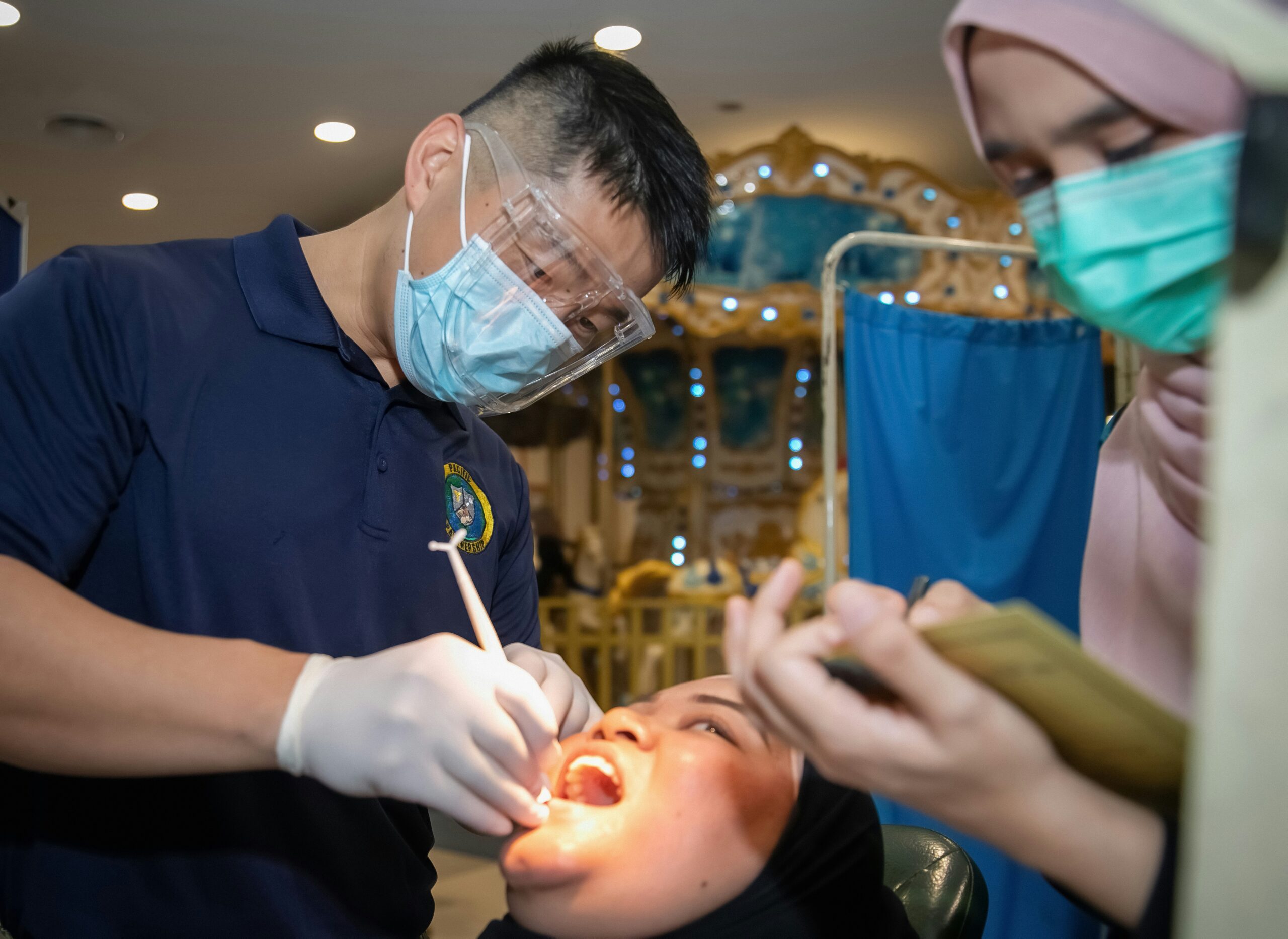 Dental professional examining patient during check-up, with another staff member taking notes, in a healthcare setting focused on patient care and accessibility.