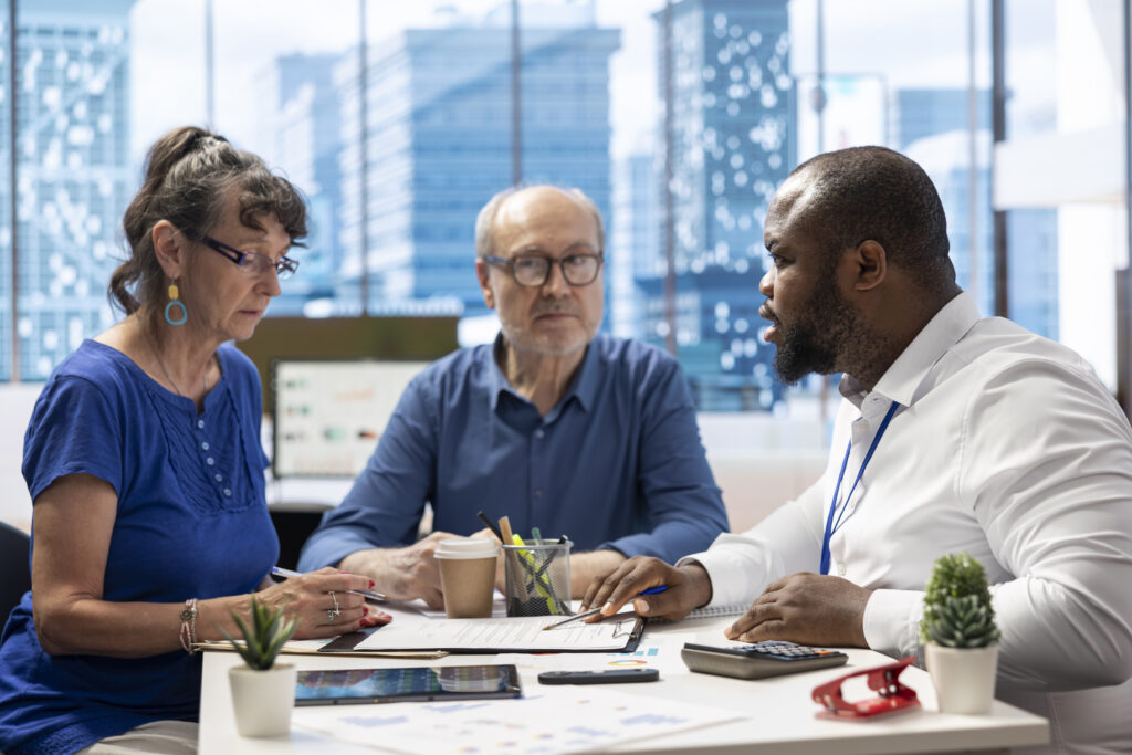 Three professionals collaborate at a conference table reviewing documents in a bright office with a cityscape outside the window.