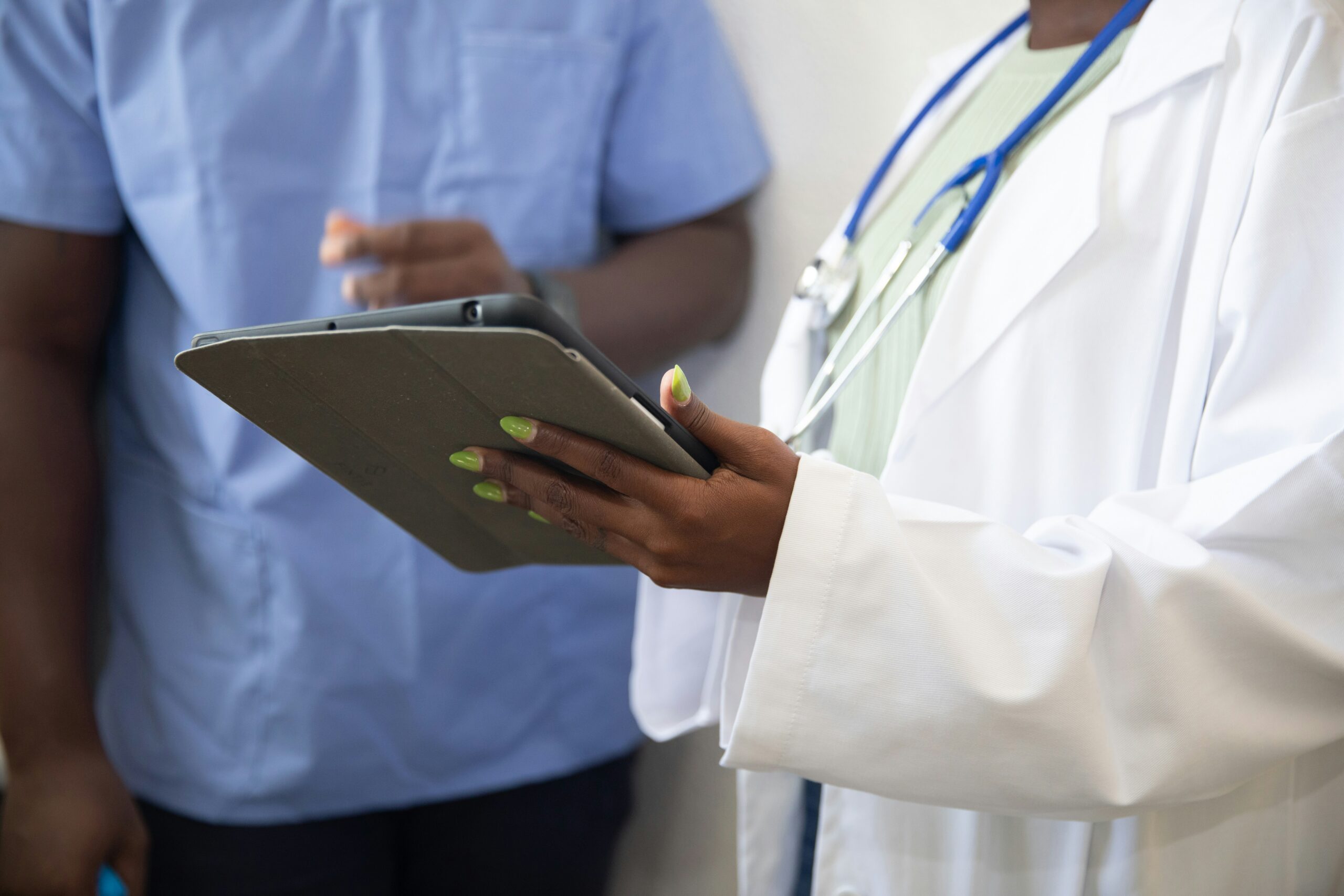 A clinician in a white coat holding a tablet while a person in blue scrubs stands nearby in a clinical setting, showing teamwork.