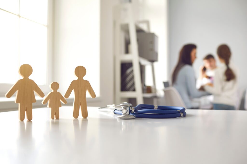 Wooden family figurines (parents and child) on a clinic desk with a stethoscope nearby, blurred clinicians in the background.