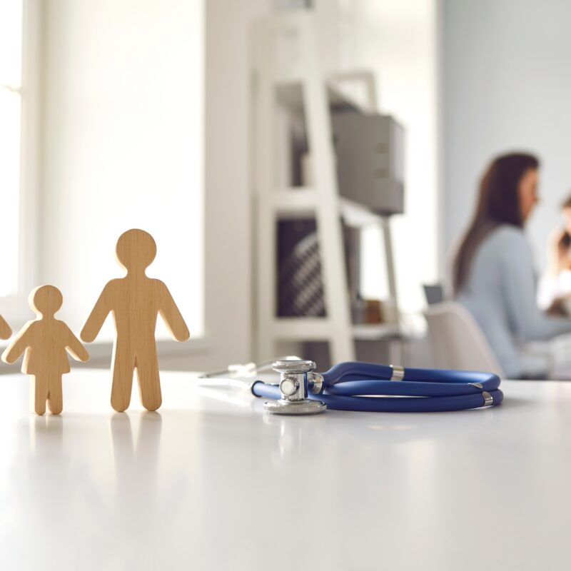 Wooden family figurines (parents and child) on a clinic desk with a stethoscope nearby, blurred clinicians in the background.