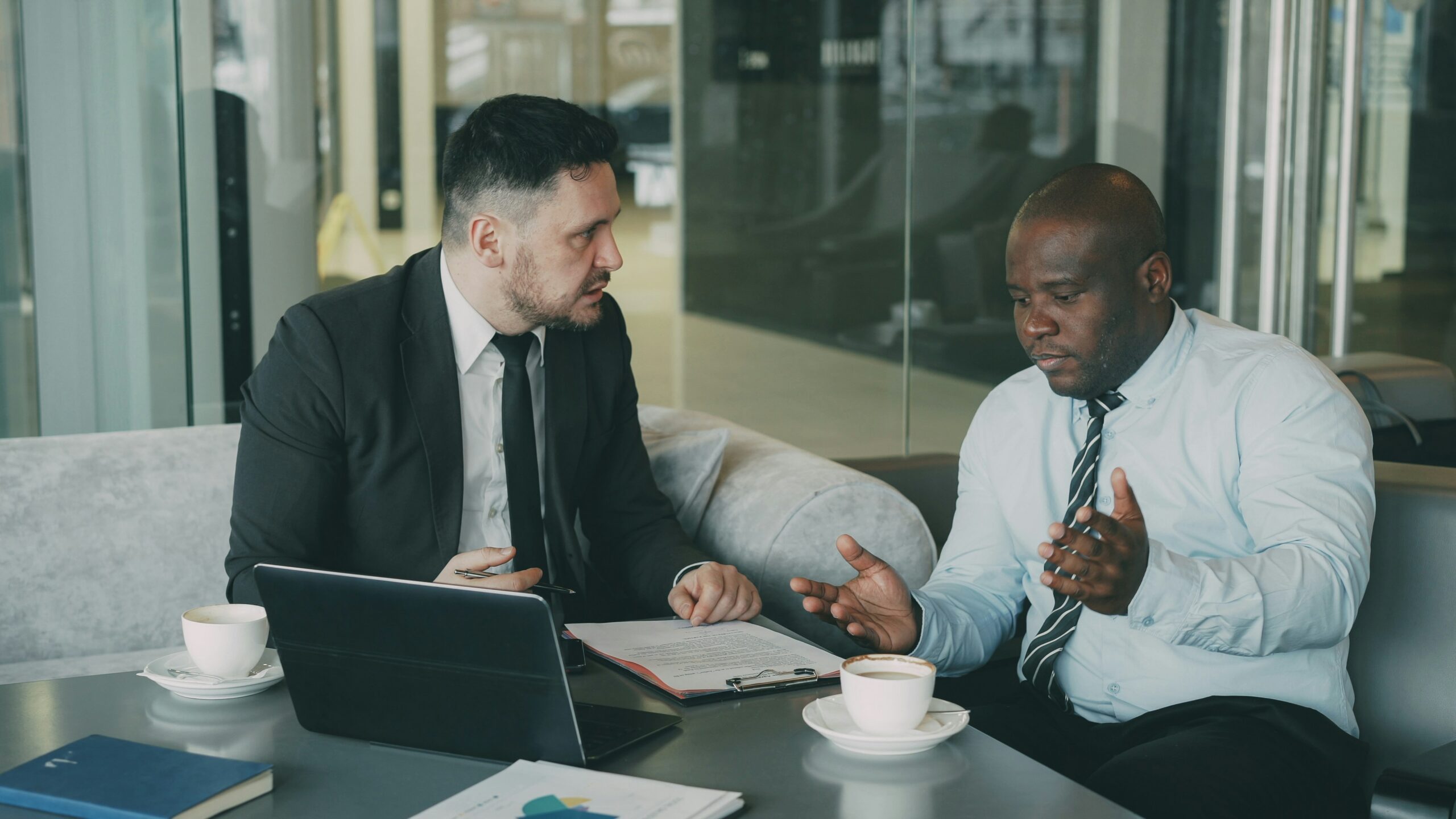 Two businessmen in a glass-walled office discuss documents at a table with coffee cups nearby.