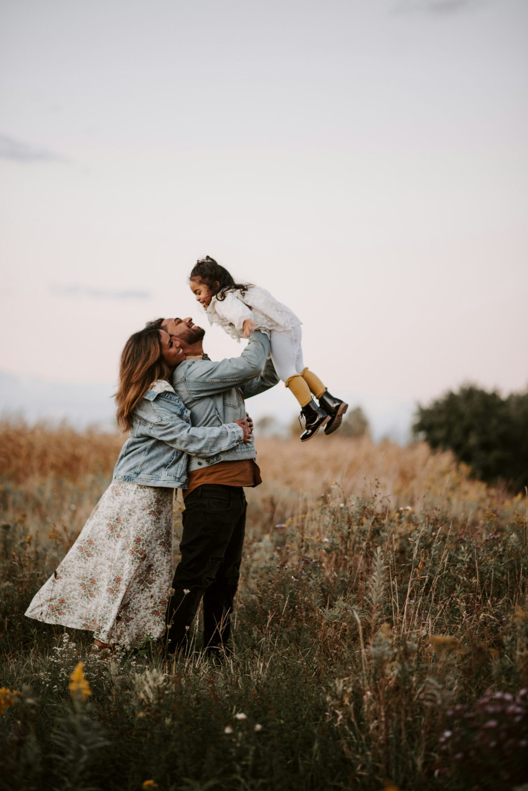 Family moment in a sunlit field: a man lifts a smiling girl as a woman looks on, all in casual clothes.