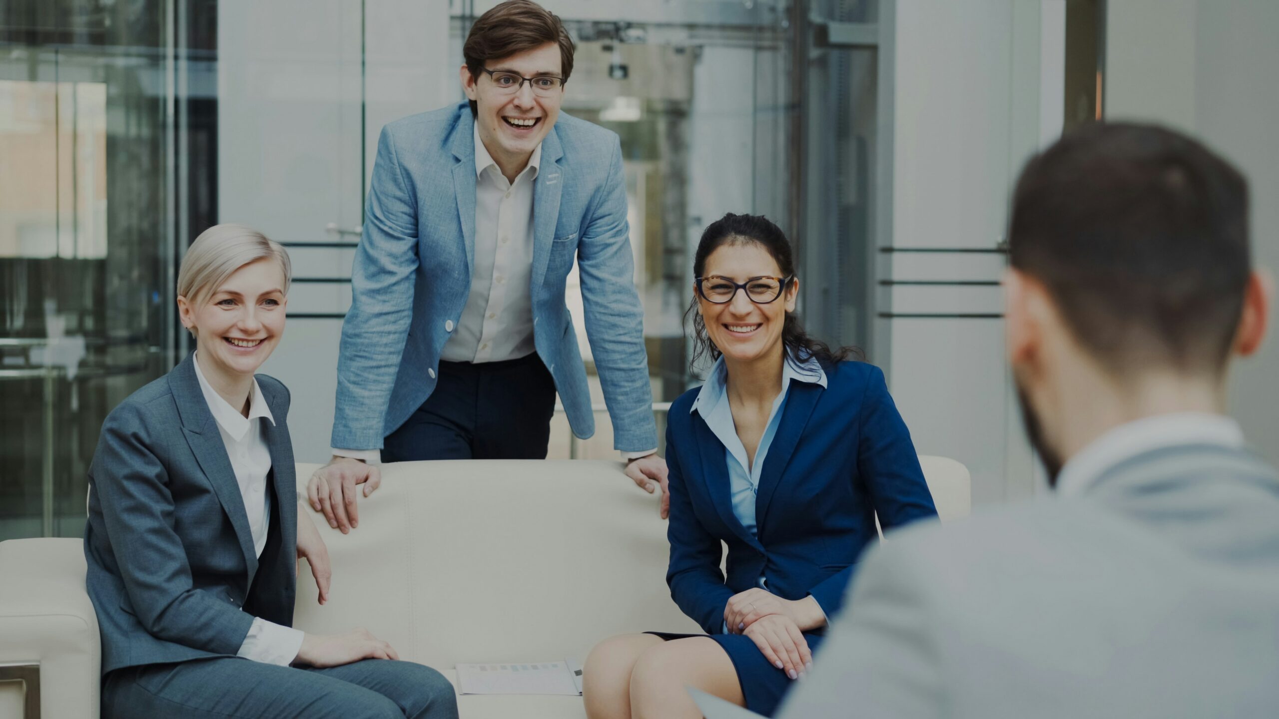 Four business professionals in a modern office, smiling and chatting around a sofa in a meeting.